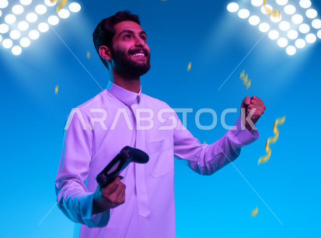 قضاء وقت ممتع في الألعاب الالكترونية والأجهزة الحديثة، العالم الرقمي الترفيهي، المتعة والتسلية بالفيديو جيمز، صورة مقربة لشاب عربي خليجي سعودي يرتدي الثوب التقليدي يمسك عصا التحكم بإيماءات وتعابير الفوز والحماس، خلفية زرقاء مع إضاءة الملعب