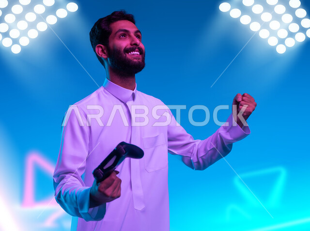 الاستمتاع في الألعاب الالكترونية والأجهزة الحديثة، العالم الرقمي الترفيهي، المتعة والتسلية بالفيديو جيمز، صورة مقربة لشاب عربي خليجي سعودي يرتدي الثوب التقليدي يمسك عصا التحكم بإيماءات وتعابير الفوز والحماس، خلفية إضاءة الملعب