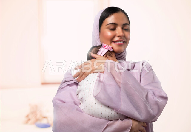 مفهوم حنان الأمهات، تحمل المسؤولية والعناية بالأطفال، قضاء وقت ممتع مع البنات، صورة مقربة لأم عربية خليجية سعودية محجبة مبتسمة تحمل طفلتها الرضيعة تمسك يدها وتحضنها بعطف وحب، مشاعر الأمومة والطفولة، خلفية وردية