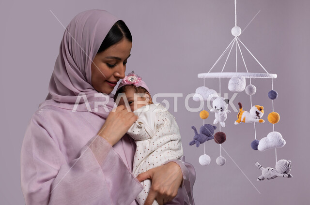 مشاعر الأمومة والطفولة، مفهوم حنان الأمهات، تحمل المسؤولية والعناية بالأطفال، قضاء وقت ممتع مع البنات، صورة مقربة لأم عربية خليجية سعودية محجبة مبتسمة تحمل طفلتها الرضيعة تمسك يدها وتحضنها بعطف وحب، خلفية رمادية مع تعليقة ألعاب