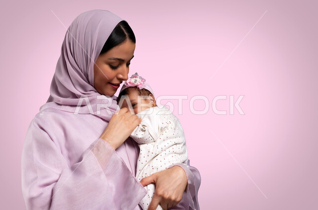 مفهوم حنان الأمهات، تحمل المسؤولية والعناية بالأطفال، قضاء وقت ممتع مع البنات، صورة مقربة لأم عربية خليجية سعودية محجبة مبتسمة تحمل طفلتها الرضيعة تمسك يدها وتحضنها بعطف وحب، مشاعر الأمومة والطفولة، خلفية وردية