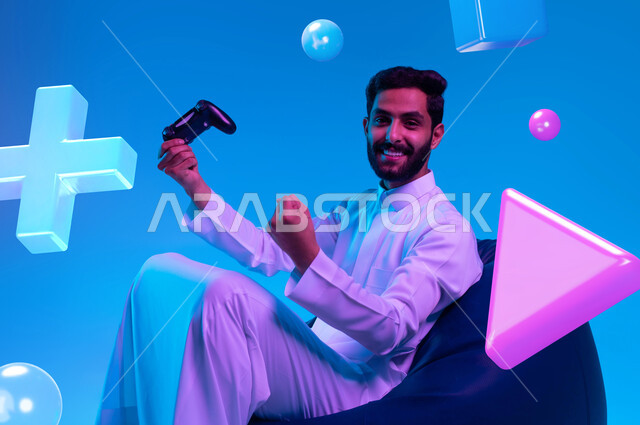 المتعة والتسلية بالفيديو جيمز، قضاء أوقات الفراغ في اللعب بالألعاب الالكترونية الترفيهية، بورتريه لشاب عربي خليجي سعودي مبتسم يرتدي الثوب التقليدي يجلس على كرسي مريح و يمسك بيده عصا التحكم ويرفع قبضته للأعلى بإيماءات الفوز، النظر إلى الكاميرا، خلفية زرقاء بأيقونات أزرار التنقل