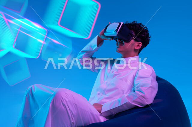 التفاعل والاندماج ومحاكاة العالم الخيالي, التسلية والترفيه والمتعه في أوقات الفراغ, استخدام التقنيات الحديثه المتطورة، بورتريه لرجل عربي خليجي سعودي يرتدي الزي التقليدي ونظارة الواقع الافتراضي يجلس على كرسي بين باج ويشاهد الافلام المفضلة, خلفية مكعبات ثلاثية الابعاد زرقاء