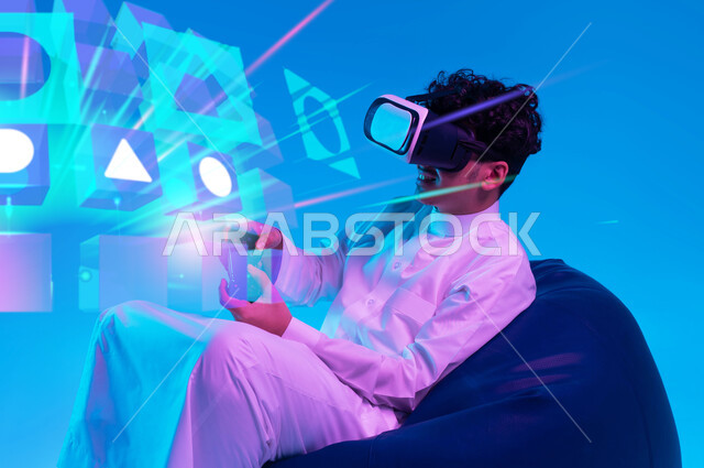 دمج التسلية مع الواقع الافتراضي والخيالي، استخدام الأجهزة التكنولوجية والتقنيات الحديثة، بورتريه لشاب عربي خليجي سعودي يرتدي الثوب التقليدي ونظارة في آر يجلس على كرسي بين باج و يمسك يد التحكم يقوم باللعب في ألعاب الفيديو الترفيهية، خلفية أيقونات أزرار التنقل ثلاثية الأبعاد