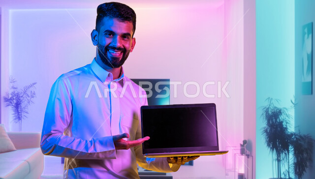 Pointing to the laptop with gestures of pleasure at modern and advanced technologies, a close-up image of a Saudi Gulf Arab man wearing a traditional dress holding a laptop, professional computer and information technology jobs, a throne of a blank black screen, living room background