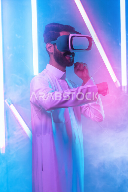 التعلم على الحركات القتالية من خلال ألعاب الفيديو، تفريغ الطاقة السلبية، ممارسة رياضة الدفاع عن النفس بتقنيات تكنولوجية، شاب عربي خليجي سعودي يرتدي الثوب التقليدي ونظارات الواقع الافتراضي بإيماءات الملاكمة والمصارعة، خلفية إنارة ملونة