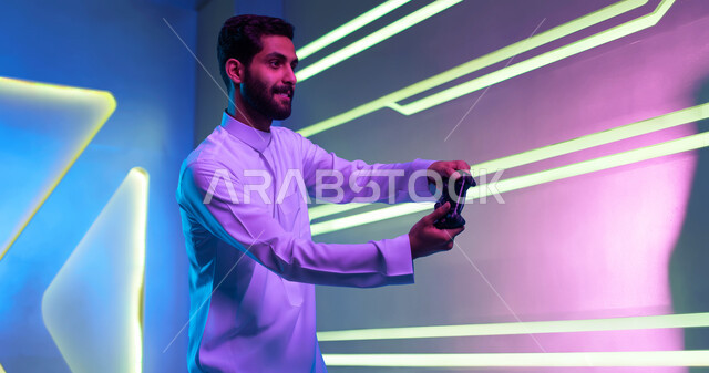 تجربة ترفيهية ممتعة ومثيرة في الألعاب الالكترونية والأجهزة الحديثة، العالم الرقمي الترفيهي، المتعة والتسلية بالفيديو جيمز، شاب عربي خليجي سعودي يرتدي الثوب التقليدي يمسك بيده عصا التحكم بإيماءات التفاعل والاندماج، خلفية إنارة ملونة