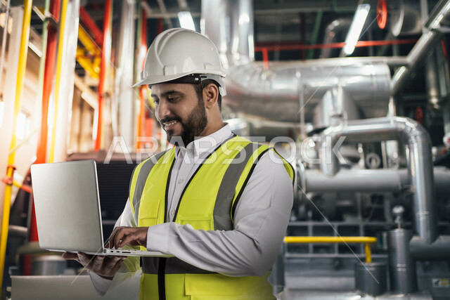 متابعة سير العملية الإنتاجية في المصانع, التطور في الإنتاج والتصنيع وتحقيق الثورة الصناعية, التحول التقني والتكنولوجي الحديث, اعتماد التشغيل الآلي و الذكاء الصنعي في الهندسة, صورة مقربة لمهندس عربي سعودي خليجي يرتدي سترة الحماية والخوذة يقوم باستخدام اللابتوب