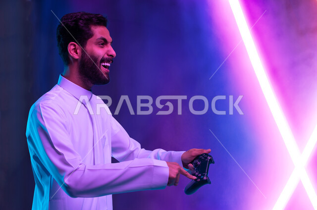 العالم الرقمي الترفيهي، تجربة ترفيهية ممتعة ومثيرة في الألعاب الالكترونية والأجهزة الحديثة، المتعة والتسلية بالفيديو جيمز، بورتريه مقرب لشاب عربي خليجي سعودي يرتدي الثوب التقليدي يمسك بيده عصا التحكم بإيماءات التفاعل والاندماج والسعادة، خلفية إنارة ملونة
