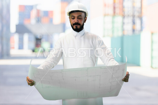 Working in the engineering sector, the concept of engineering and construction, supervising the implementation of architectural plans and projects in Saudi Arabia, a smiling Saudi Gulf Arab engineer with a protection helmet holding a construction plan in his hand.