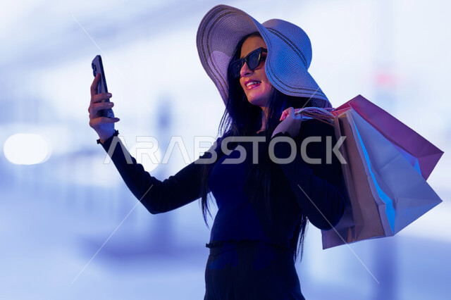 استخدام الجوال في التسوق عبر الانترنت، صورة مقربة لامرأة عربية خليجية سعودية ترتدي النظارة الشمسية تحمل أكياس ورقية ملونة تتسوق في المركز التجاري تمسك بيدها الهاتف المحمول, ازياء واكسسوارات نسائية، التقاط صور السيلفي التذكارية