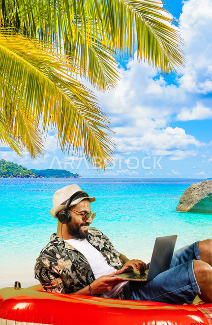 الترفيه والتسلية بالعطلة الصيفية, الرحلات السياحية في شواطئ المملكة, شاب عربي خليجي سعودي يرتدي الزي الكاجوال وسماعة الهيدفون يستخدم الحاسب المحمول, التواصل مع الأصدقاء عبر الأونلاين, الاستمتاع بسماع المقاطع الموسيقية والتسجيلات الصوتية, خلفية شاطئ البحر