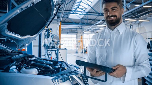 اكتشاف الأعطال وتصليح السيارات في ورشة الصيانة، مهندس ميكانيكي محترف ينظر إلى الكاميرا بإيماءات الثقة يقوم بتصليح عطل السيارة عبر جهاز التابليت، استخدام الأجهزة والتقنيات الحديثة والمتطورة, خلفية ورشة العمل