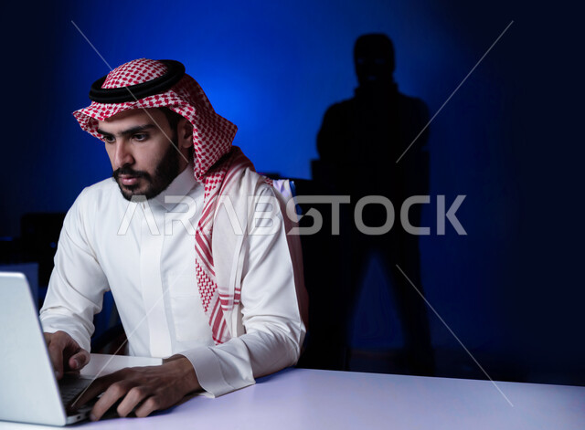 التعرض للهجوم التقني, صورة مقربة لشاب عربي خليجي سعودي يرتدي الثوب التقليدي والشماغ يستخدم الحاسب المحمول, اختراق حماية وأمن وخصوصية المستخدمين, مفهوم الحفاظ على نظام الأمن الالكتروني, خلفية رجل هاكر ملثم