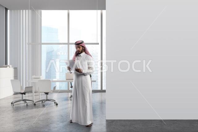 متابعة الأهداف والخطط في مقر الشركة, رجل عربي خليجي سعودي يرتدي الثوب التقليدي والشماغ يمسك بيده أوراق المشاريع, العمل في قطاع المكاتب والشركات السعودية, بيئة العمل المكتبية, قراءة البنود الخاصة بالعقود والاتفاقيات, التحقق والتدقيق في الشروط