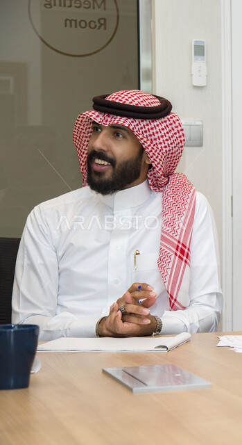 اجتماع عمل داخل مقر الشركة, رجل أعمال عربي خليجي سعودي مبتسم يرتدي الثوب التقليدي والشماغ يناقش الأفكار الجديدة بإيماءات الثقة والسرور, مهن ووظائف مكتبية, نظام العمل في المكاتب والشركات السعودية, الالتزام بإنجاز المهام