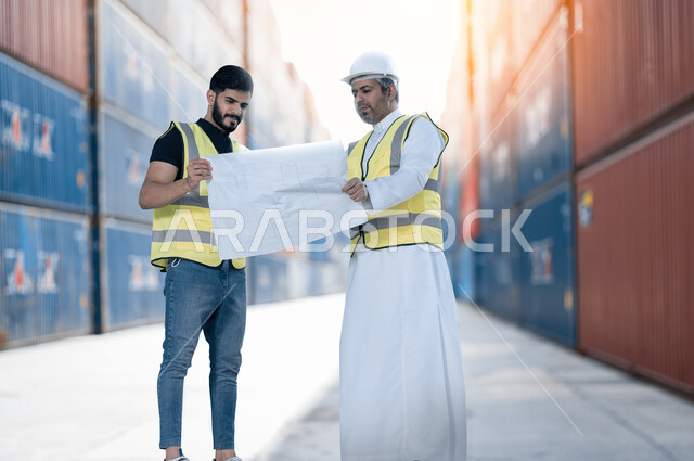 إعطاء التعليمات من قبل المدير, مهندسان عربيان سعوديان خليجيان يرتديان سترة الحماية يمسكان مخطط المشروع، الإستيراد والتصدير في السعودية, إدارة الأعمال والإشراف على سير الخطة الهندسية والتدقيق على إنجاز المهام, خلفية موقع العمل