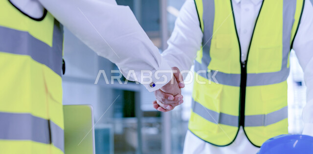 عقد صفقات جديدة, اتفاقية عمل ناجحة, صورة مقربة لمهندسين عربين خليجيين سعوديين يرتديان سترة الحماية يقومان بالتصافح والسلام في مقر الشركة, العمل في القطاع الهندسي, مهن ووظائف سعودية, تحقيق أهداف الشؤون الهندسية