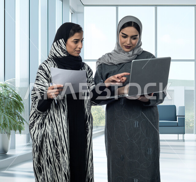 Professions and jobs for women, developing strategies and exchanging ideas and experiences to achieve work goals, cooperation and team spirit, two Saudi and Gulf Arab female colleagues discuss and organize business using the laptop, integrating work with technology and technology.