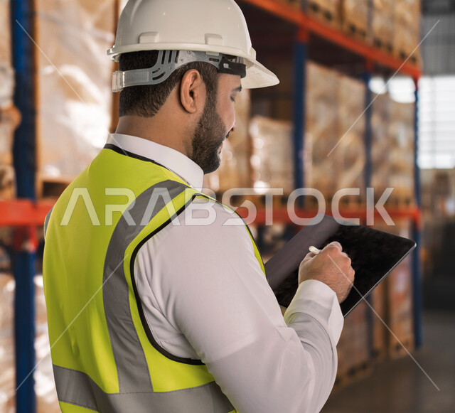 مفهوم الهندسة والانشاءات، العمل في القطاع الهندسي، استخدام الأجهزة والتقنيات الحديثة في الأعمال الهندسية, صورة مقربة من الجانب لمهندس عربي خليجي سعودي يرتدي السترة وخوذة الحماية يمسك بيده جهاز التابليت, الإشراف على خطة الانتاج ومتابعة شؤون المصنع