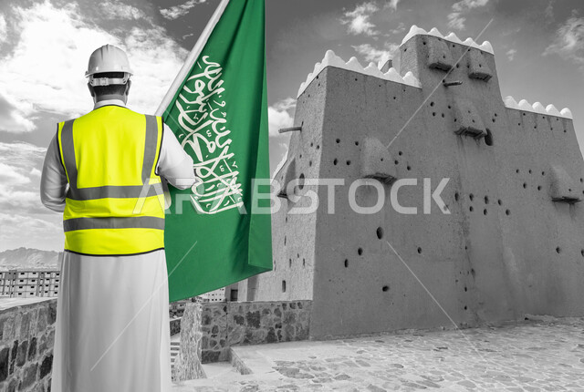 إحياء ذكرى اليوم الوطني السعودي في 23 سبتمبر, دور المهندسيين في التطور العمراني, الاحتفال بيوم العلم 11 مارس, صورة من الخلف لمهندس عربي سعودي خليجي يرتدي سترة الحماية والخوذة البيضاء يمسك علم المملكة بفخر واعتزاز, خلفية الأماكن الأثرية والطينية