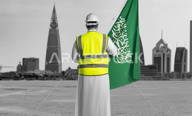 دور المهندسيين في التطور العمراني, إحياء ذكرى اليوم الوطني السعودي في 23 سبتمبر, الاحتفال بيوم العلم 11 مارس, صورة من الخلف لمهندس عربي سعودي خليجي يرتدي سترة الحماية والخوذة البيضاء يمسك علم المملكة بفخر واعتزاز, خلفية الأبراج وناطحات السحاب المشهورة