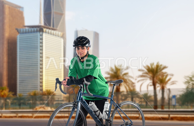 أنشطة ورضيات نسائية، مواصلات آمنة في المملكة، جودة الحياة الصحية، شابة عربية خليجية سعودية ترتدي الملابس الرياضية وخوذة الحماية تتجول على الدراجة الهوائية في الهواء الطلق، ممارسة نشاط بدني للمحافظة على اللياقة الجسدية، خلفية الأبراج وناطحات السحاب المشهورة