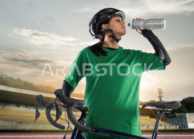 جودة الحياة الصحية، نشاط بدني للمحافظة على اللياقة الجسدية، شابة عربية خليجية سعودية مبتسمة ترتدي الملابس الرياضية وخوذة الحماية تقوم بشرب الماء أثناء ممارسة رياضة ركوب الدراجة الهوائية في الهواء الطلق، أنشطة ورياضات نسائية في المملكة