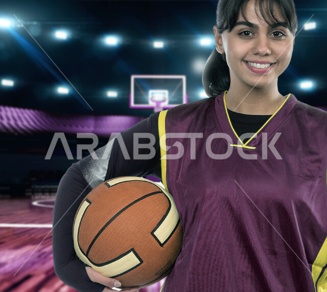 النظر إلى الكاميرا بإيماءات السعادة والسرور، أنشطة ورياضيات نسائية في المملكة، شابة عربية خليجية سعودية مبتسمة ترتدي الملابس الرياضية تحمل بيدها كرة السلة في الملعب، تمارين بدنية للتسليه والترفيه، جودة الحياة الصحية، مفهوم اللياقة والمرونة