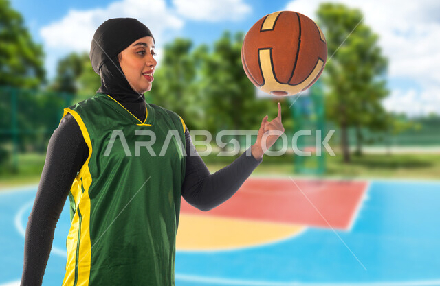 أنشطة ترفيهية ورياضات نسائية، التدرب على اتقان لعب كرة السلة في الملعب، إجراء تمرينات رياضية في الهواء الطلق، لاعبة عربية خليجية سعودية محجبة ترتدي ملابس رياضية تقوم بموزانة الكرة عى اصبعها، إيماءات وتعابير التركيز والانتباه، نمط الحياة الصحية