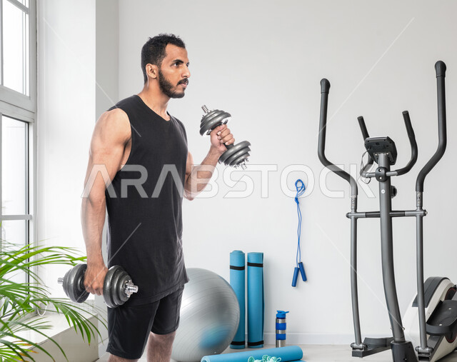 نشاط بدني للمحافظة على الصحة واللياقة الجسدية، رفع الأثقال والأوزان الحديدية الثقيلة، شاب عربي خليجي سعودي يمارس التمارين الرياضية مع الدمبلز، كمال الأجسام وتقوية وبناء العضلات، أسلوب حياة صحي، خلفية النادي الرياضي
