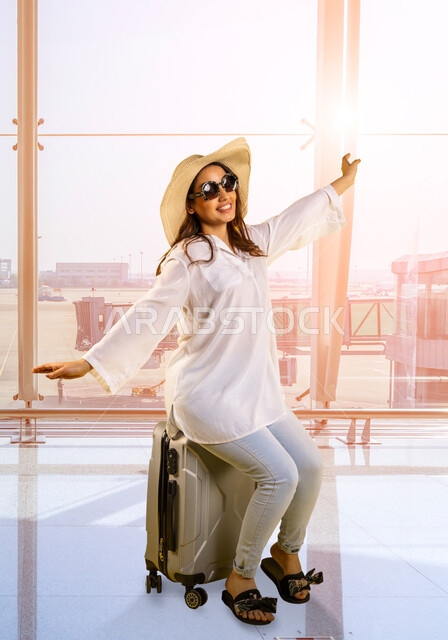 Spending fun times traveling outside the country, a Saudi Gulf Arab woman wearing a casual outfit and a straw hat with sunglasses sitting on a suitcase with expressions and gestures of enthusiasm and pleasure, enjoying summer trips, airport background