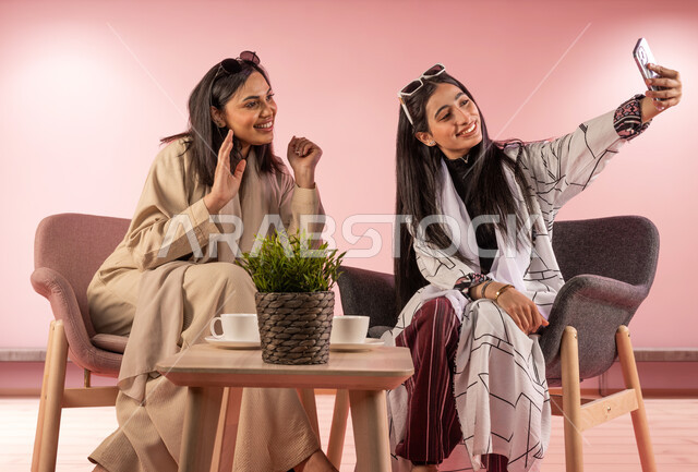 توثيق اللحظات السعيدة باستخدام الهاتف المحمول، بورتريه لفتاة عربية خليجية سعودية تقوم بالتقاط صورة سيلفي تذكارية مع صديقتها بتعابير وايماءات السرور والسعادة، قضاء اوقات ممتعة برفقة الاصدقاء، جلسات وناسة نسائية، خلفية وردية