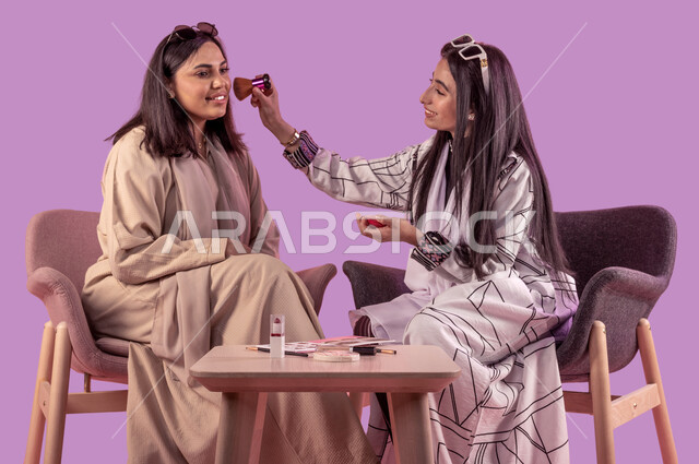 التجهز والاستعداد للذهاب مع الاصدقاء، إستخدام ادوات الميكاب تتوريال، خبيرة تجميل محترفة، بورتريه لشابة عربية خليجية سعودية تقوم بوضع المكياج وتزين صديقتها بتعابير وايماءات السرور، الاهتمام بالأناقة والمظهر الخارجي، خلفية بنفسجية