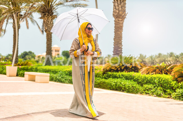 Walking and picnicking in one of the Kingdom’s recreational parks during the day, a veiled Saudi Arabian Gulf woman wearing a modern abaya and sunglasses, holding an umbrella in her hand, enjoying the green park.