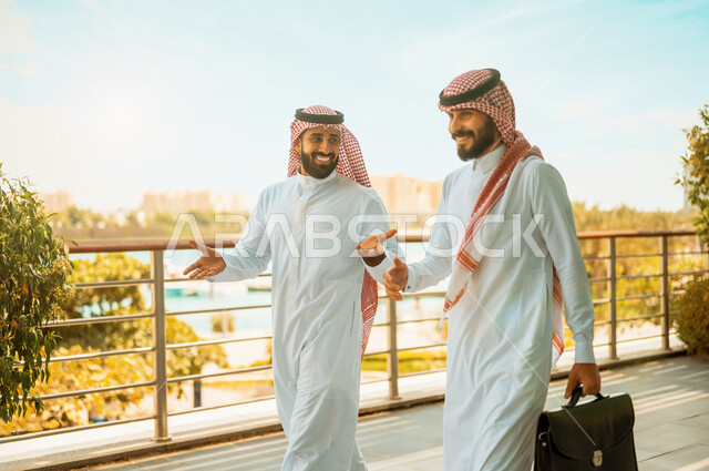 Friends and co-workers, the end of work and the departure of employees, two Saudi-Gulf Arab businessmen wearing the shemagh and traditional thobe discussing with happy gestures, Saudi professions and jobs, discussions and dialogues outside the company’s headquarters, successful business partnerships