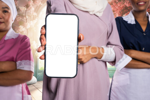 موك آب أبيض مستطيل، صورة مقربة لسيدة عربية خليجية سعودية تمسك بيدها الهاتف المحمول بشاشة بيضاء فارغة بجانبها عاملات النظافة بإيدي متقاطعة، استقدام العمالة المنزلية المؤهلة والمرخصة من الخارج، خلفية منزل بديكورات وتصاميم حديثة