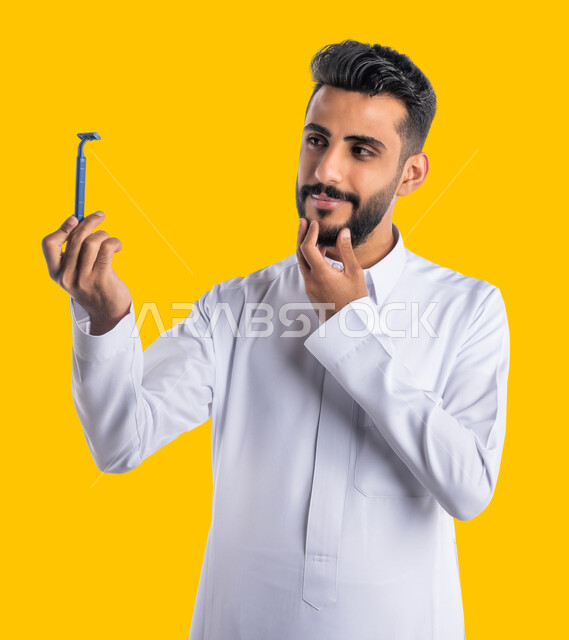 مفهوم العناية بالبشرة والاهتمام بالمظهر الخارجي، المحافظة على الأناقة والنظافة الشخصية، بورتريه مقرب لرجل عربي خليجي سعودي يرتدي الثوب التقليدي يمسك بيده شفرة الحلاقة الرجالية وينظر إليها بإيماءات الإعجاب والسرور، خلفية صفراء