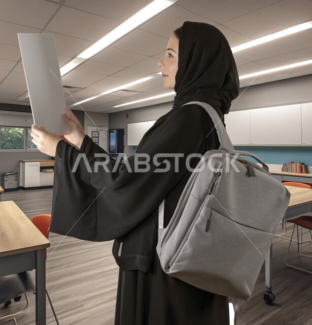 متابعة المحاضرات والدروس، مفهوم الاجتهاد والمثابرة، جودة التعليم في المدارس والجامعات السعودية، طالبة جامعية خليجية عربية سعودية مبتسمة ترتدي العباءة السوداء تحمل بيدها الحقيبة الدراسية وورة بيضاء تنظر إليها بإيماءات التركيز، خلفية الفصل الدراسي