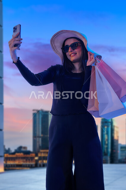 استخدام الجوال في التسوق عبر الانترنت، صورة مقربة لامرأة عربية خليجية سعودية ترتدي النظارة الشمسية وقبعة تحمل أكياس ورقية ملونة تمسك بيدها الهاتف المحمول, ازياء واكسسوارات نسائية، التقاط صور السيلفي التذكارية، خلفية الابراج والمعالم المشهورة في السعودية