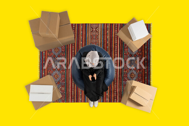 عملية شحن وتوصيل موثوقه, الدفع عن طريق البطاقه البنكيه في طلبات الاونلاين, بورتريه علوي عامودي لإمرأة عربية سعودية خليجية ترتدي العباءة السوداء تجلس على كرسي مريح تمسك الهاتف المحمول بشاشة سوداء فارغة، التسوق والشراء عن طريق الانترنت, خلفيه صفراء