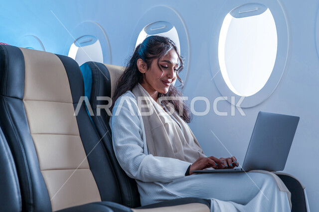 مشاهدة الأفلام والمسلسلات، إنجاز مهام العمل عن بعد, ايماءات تدل على السعادة والسرور, شابة عربية سعودية خليجية مبتسمة ترتدي الزي الكاجوال تجلس على مقعد في الطائرة تستخدم الحاسوب المحمول، وسائل النقل الجوي، الخطوط الجوية السعودية