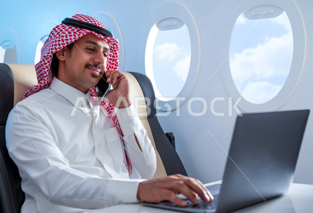 استخدام الهاتف المحمول، إنجاز مهام العمل عن بعد, ايماءات تدل على السعادة والسرور, رجل عربي سعودي خليجي مبتسم يرتدي الزي التقليدي يجلس على مقعد في الطائرة يستخدم الحاسوب المحمول يقوم بإجراء مكالمات هاتفيه، وسائل النقل الجوي، الخطوط الجوية السعودية