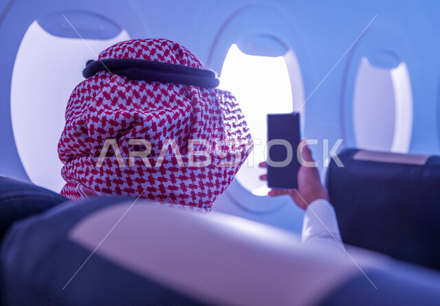 استخدام الجوال لالتقاط صور السيلفي التذكارية, صورة مقربة من الخلف لرجل عربي سعودي خليجي يرتدي الزي التقليدي يجلس على مقعد في الطائرة يستخدم الهاتف المحمول، وسائل النقل الجوي، توثيق اللحظات السعيدة، الخطوط الجوية السعودية