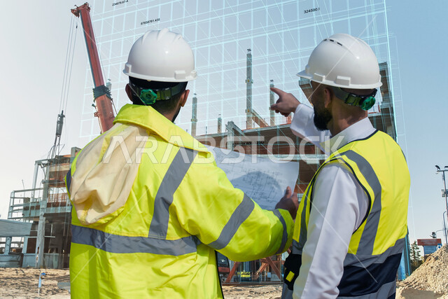 العمل في القطاع الهندسي، الحرص على بناء الأساسيات الجيده للمبنى، صورة مقربة من الخلف لمهندسان عربيان خليجيان سعوديان يرتديان الخوذة وسترة الحماية يتناقشان حول المشروع، إيماءات الإشارة إلى شيء ما، التحقق من تطبيق الخطط و المشاريع الهندسيه
