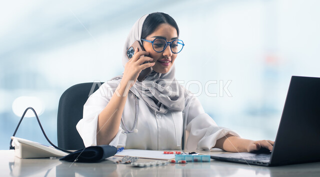 تقديم الخدمات الطبية عن بعد، استشارات صحية عبر الجوال، صورة مقربة لطبيبة عربية خليجية سعودية مبتسمة ترتدي الحجاب والمعطف الابيض والنظارات الطبية تقوم بالعمل على الحاسب المحمول، مفهوم الطب والرعاية الصحية، خلفية ضبابية للعيادة