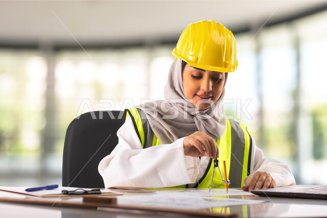 استخدام أدوات ومعدات هندسية على مخطط تصميم هندسي، انجاز وتتبع المهام عبر الحاسب المحمول، مهندسة معمارية عربية خليجية سعودية ترتدي الخوذة وسترة الحماية تجلس داخل المكتب تقوم بالرسم، مهن ووظائف سعودية، خلفية مكتبية ضبابية
