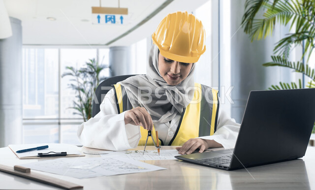 انجاز وتتبع المهام عبر الحاسوب المحمول ، استخدام أدوات ومعدات هندسية على مخطط تصميم هندسي ، مهندسة معمارية عربية خليجية سعودية ترتدي الخوذة وسترة الحماية تجلس داخل المكتب تقوم بالرسم، مهن ووظائف سعودية، خلفية مكتبية
