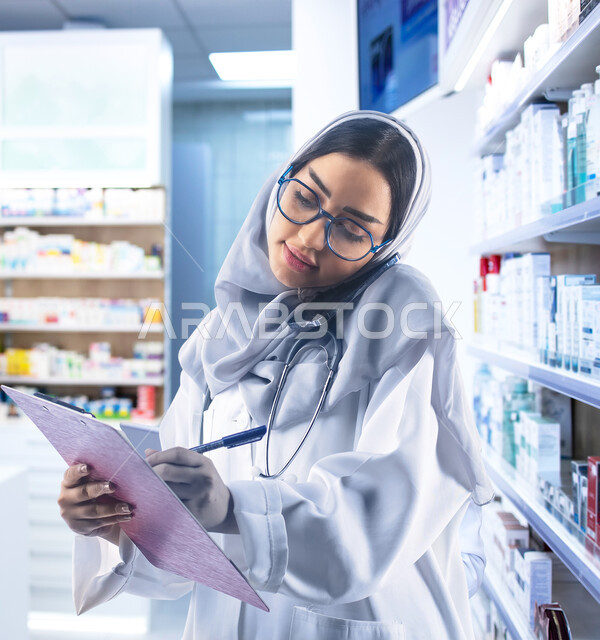 تسجيل وتدوين معلومات المريض، صورة مقربة لطبيبة صيدلانية عربية سعودية خليجية محجبة ترتدي المعطف الابيض والنظارات الطبية تضع سماعة نبضات القلب على رقبتها تقوم بإجراء مكالمة هاتفية باستخدام الجوال، تقديم الخدمات العلاجية والصحية عن بعد، خلفية الصيدلية