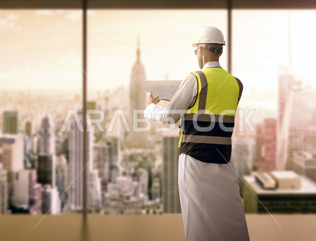 وظائف ومهن سعودية, إيماءات الافتخار بالنهضة العمرانية للمملكة, مشاريع هندسية تنموية وطنية للبلاد, صورة من الجانب لمهندس عربي سعودي خليجي يرتدي الخوذة وسترة الحماية يحمل بيده مخطط الإنشاء يقوم بمقارنته مع إنجازات الواقع, خلفية أبراج الرياض